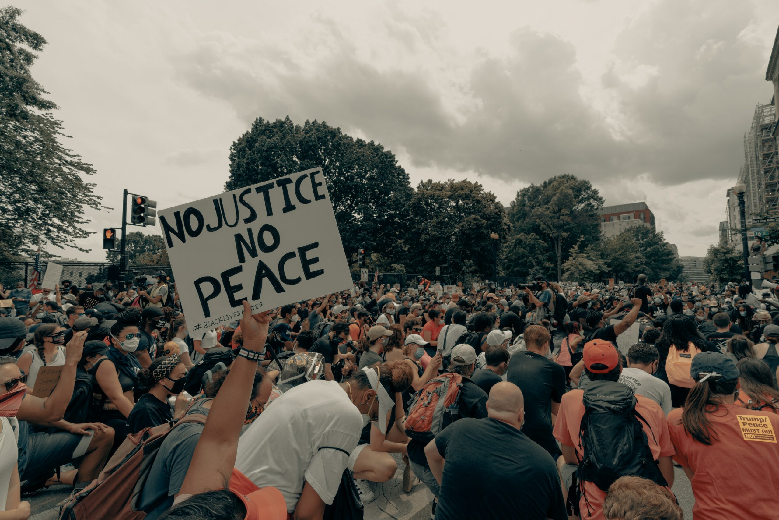 A crowd of people at a protest kneel on the ground. A prominent sign read, "No justice, no peace."